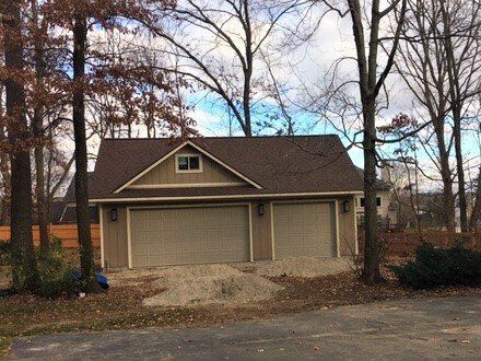 Two-car garage with beige siding and dark roof, viewed from a tree-lined street on an overcast day