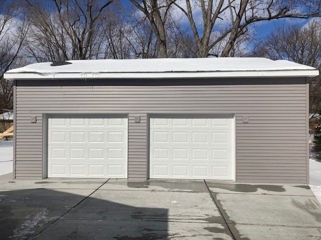 Two-car detached garage with white doors, gray siding, and a snow-covered roof in a driveway