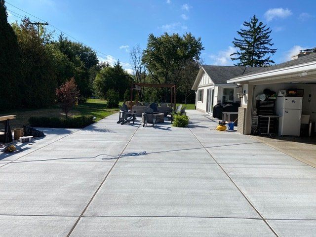 Backyard patio with concrete slab, outdoor seating, open garage, and trees under a sunny blue sky