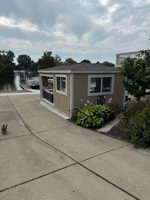 Small tan utility building beside a concrete walkway, with trees and a fountain in the background.