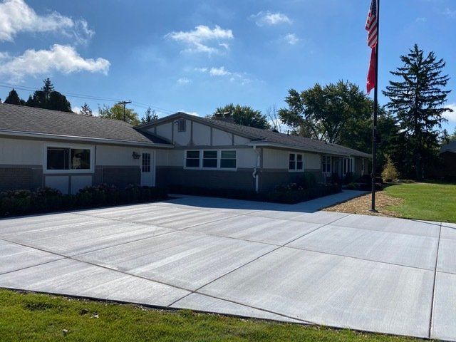 Single-story house with a wide concrete driveway, green lawn, and an American flag under a blue sky.