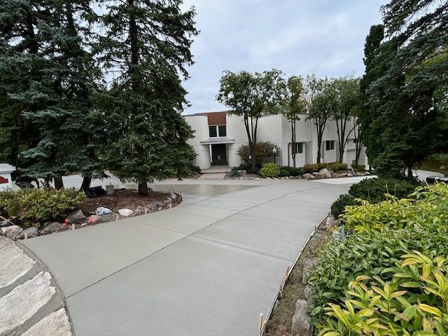 Curved concrete walkway leading to a white building entrance, lined with trees and shrubs.