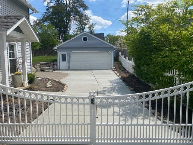 Gated driveway leading to a detached garage beside a house, with concrete paving and trees lining the side.
