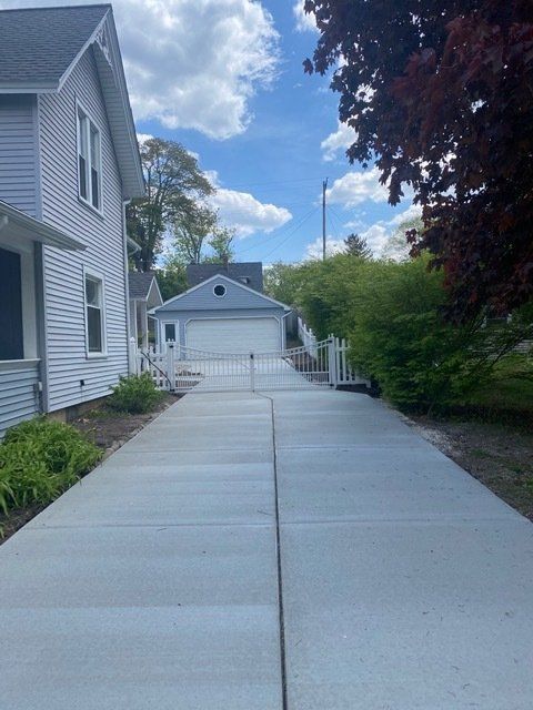 Long concrete driveway beside a gray house leading to a detached garage under a partly cloudy sky