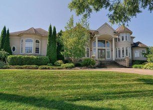 Large beige suburban house with arched windows, trees, and a wide green lawn on a sunny day