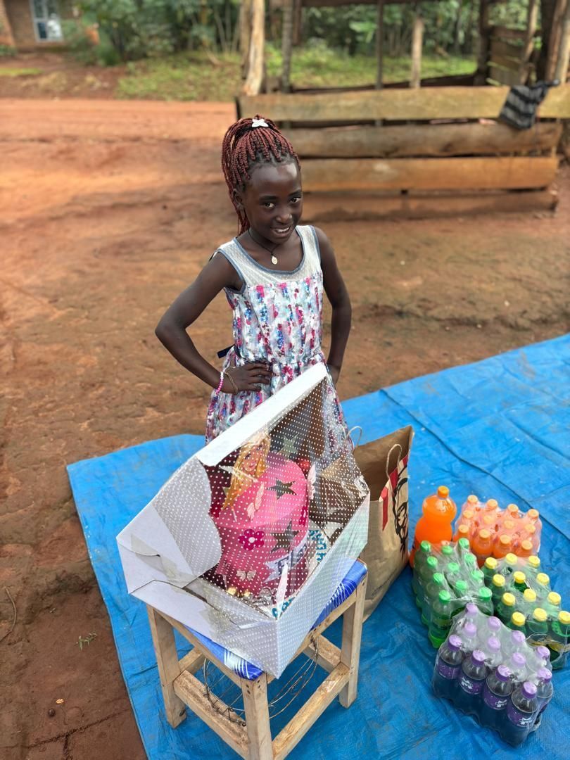 Girl with braided hair stands at a table with items for sale, like drinks. She has her hand on her hip and smiles.