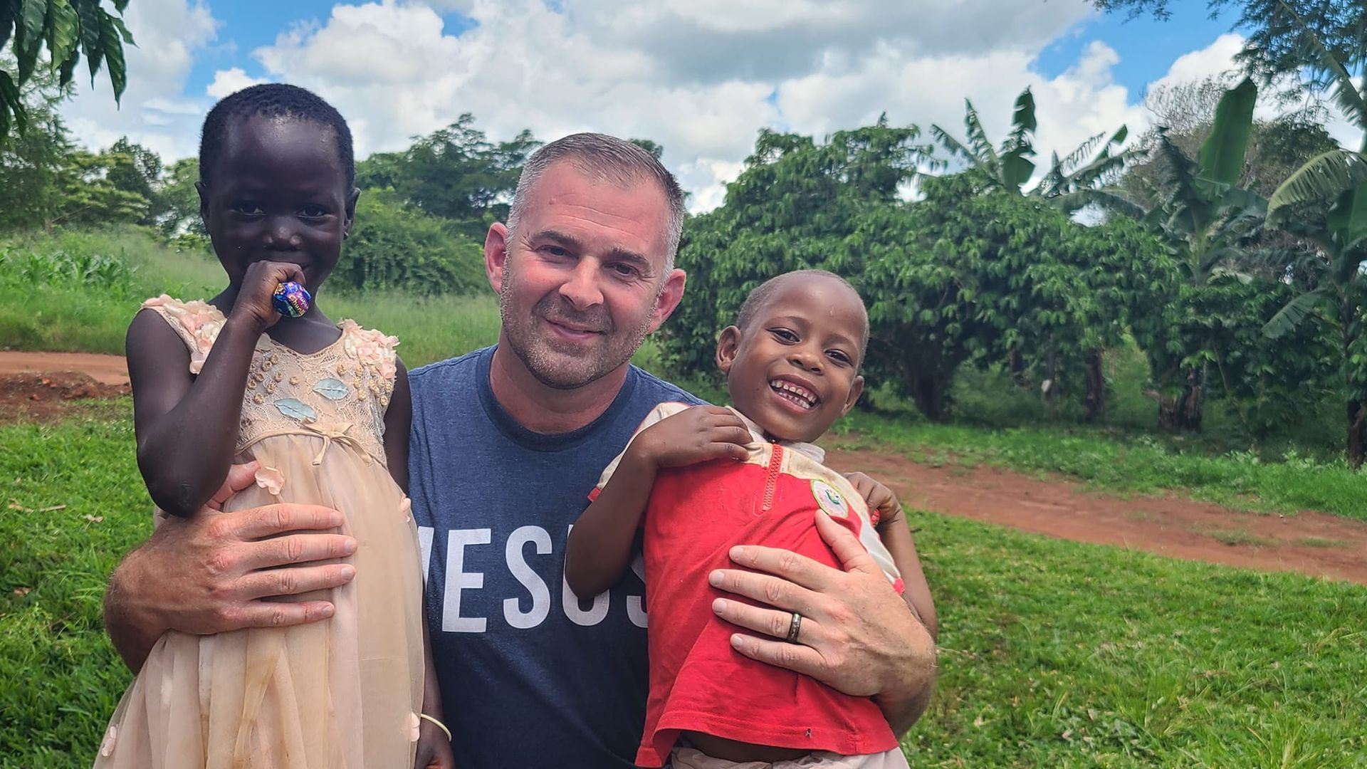 Man smiles, holding two children outdoors in a grassy area with trees; one child is holding a flower.