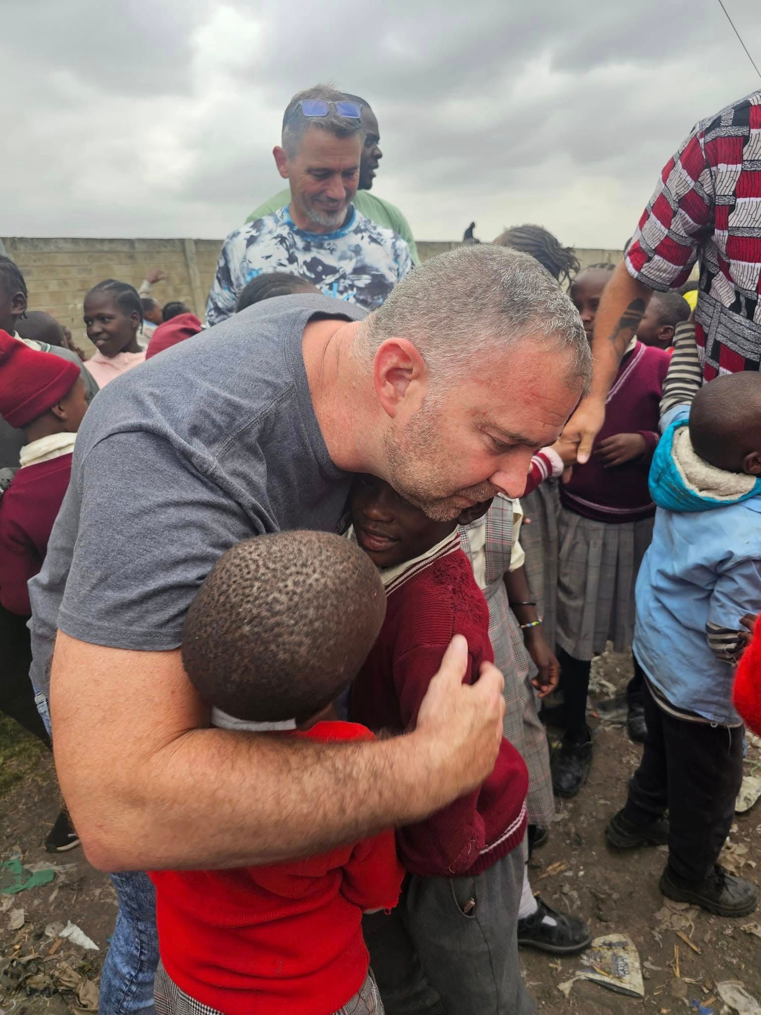 Man hugging a child, surrounded by other children in a school setting, cloudy outdoor background.