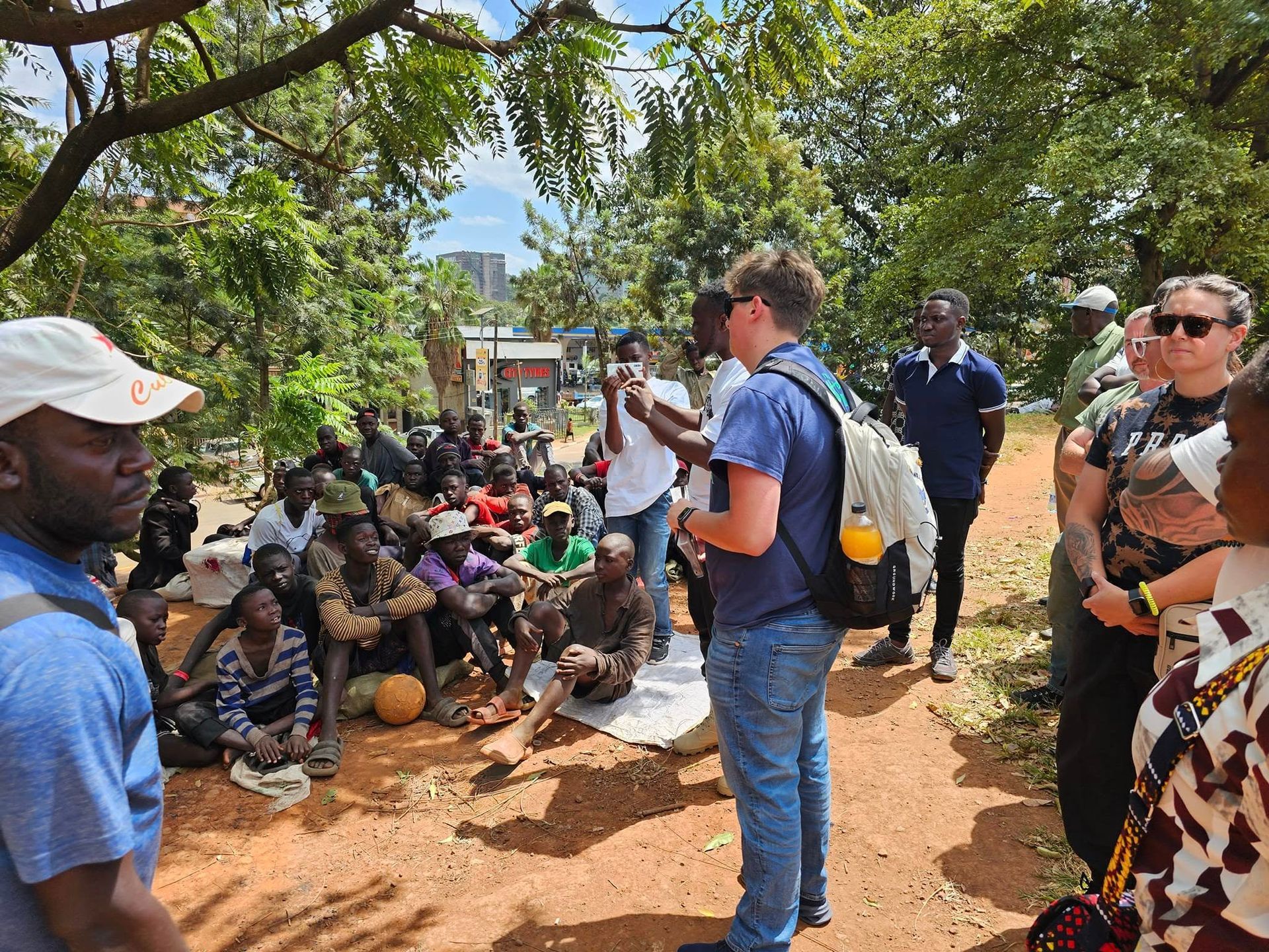 Group of people outdoors, some seated on the ground. Several individuals in foreground, with trees and buildings visible.