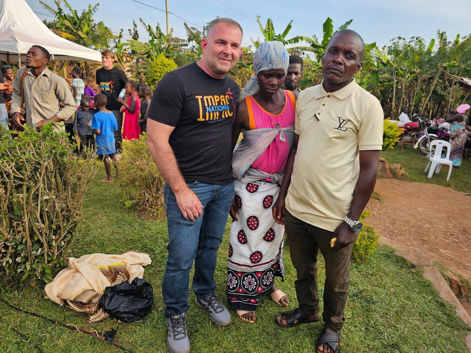 Three people pose outdoors; a man in black t-shirt, a woman in pink dress, and a man in a yellow shirt. People are in the background.