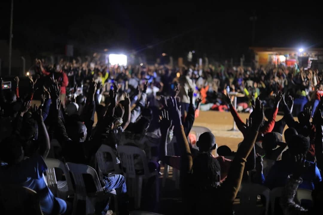 Large crowd outdoors at night with arms raised. Bright lights illuminate the stage area.