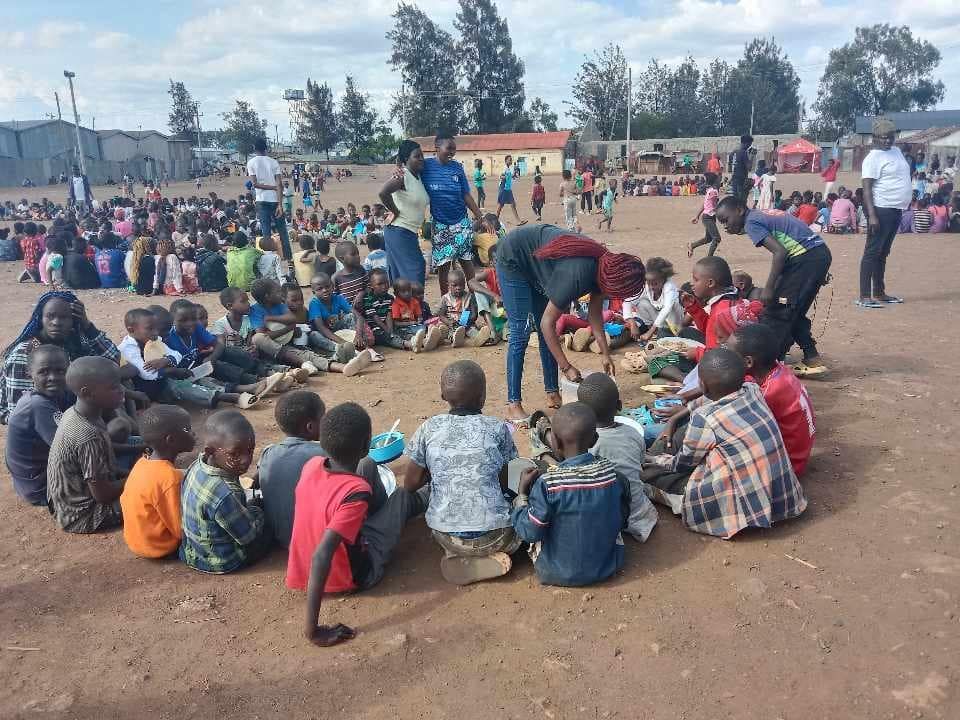 Children and adults seated in a circle on a dirt field; a group activity or event taking place outdoors.
