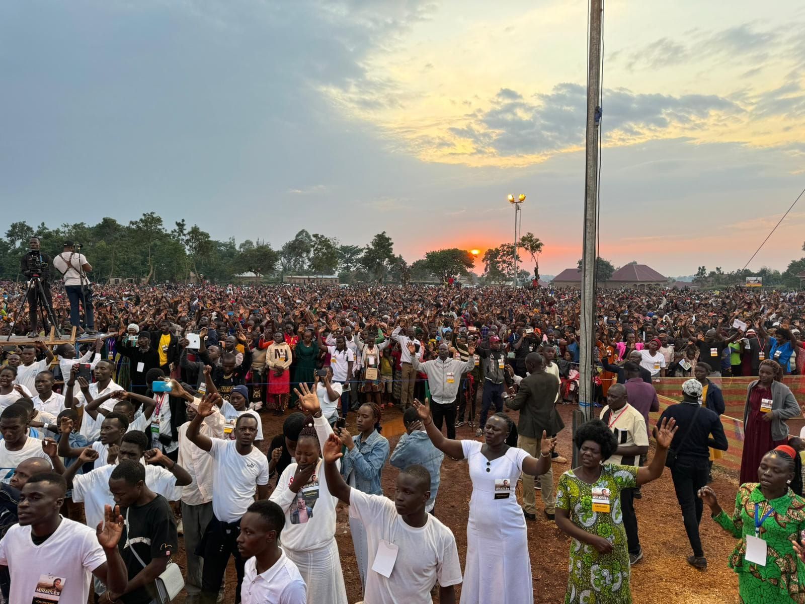 Large crowd outdoors with arms raised, likely at a religious event, under a cloudy sky.