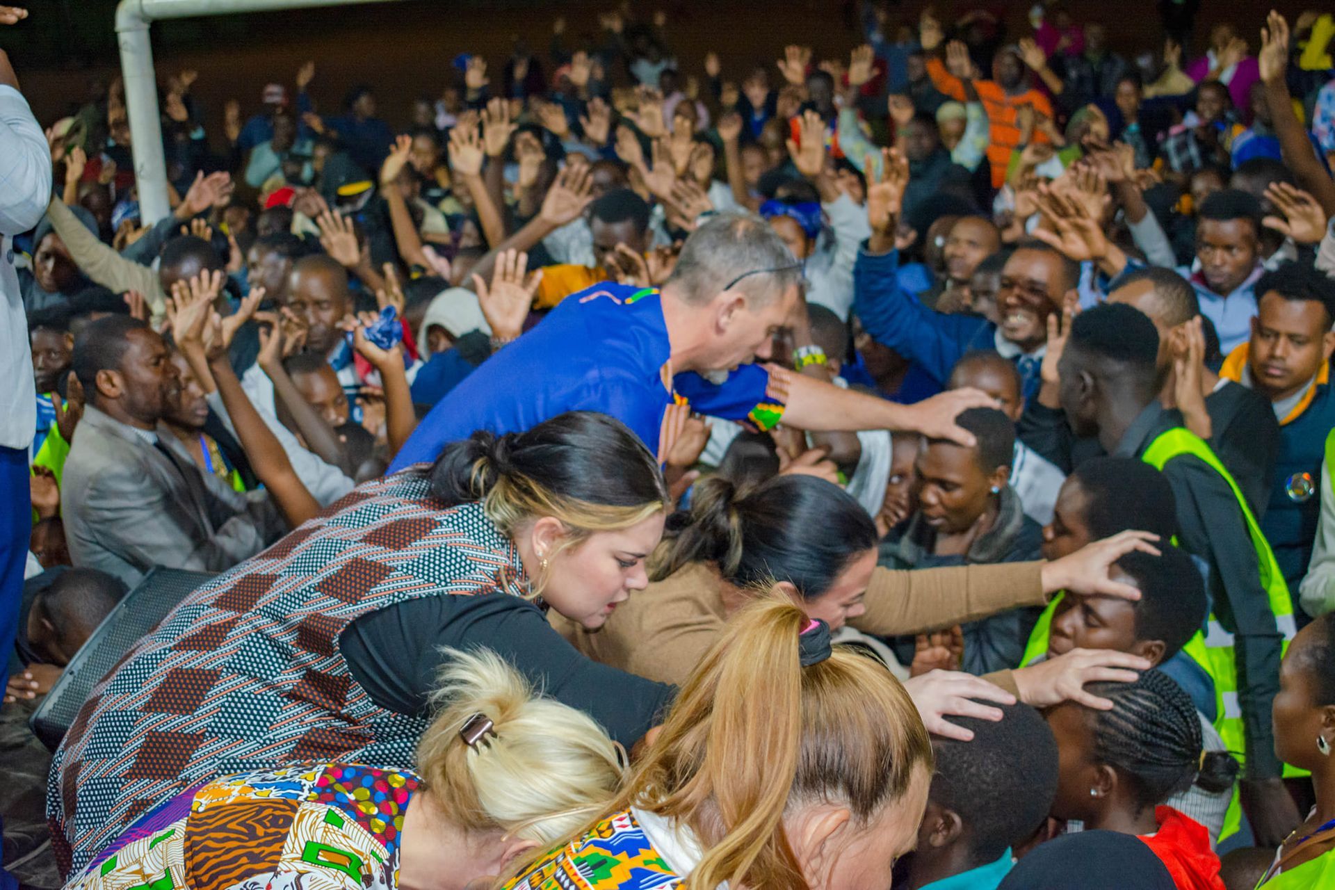 People reaching toward others with raised hands; group praying, outdoors at night.