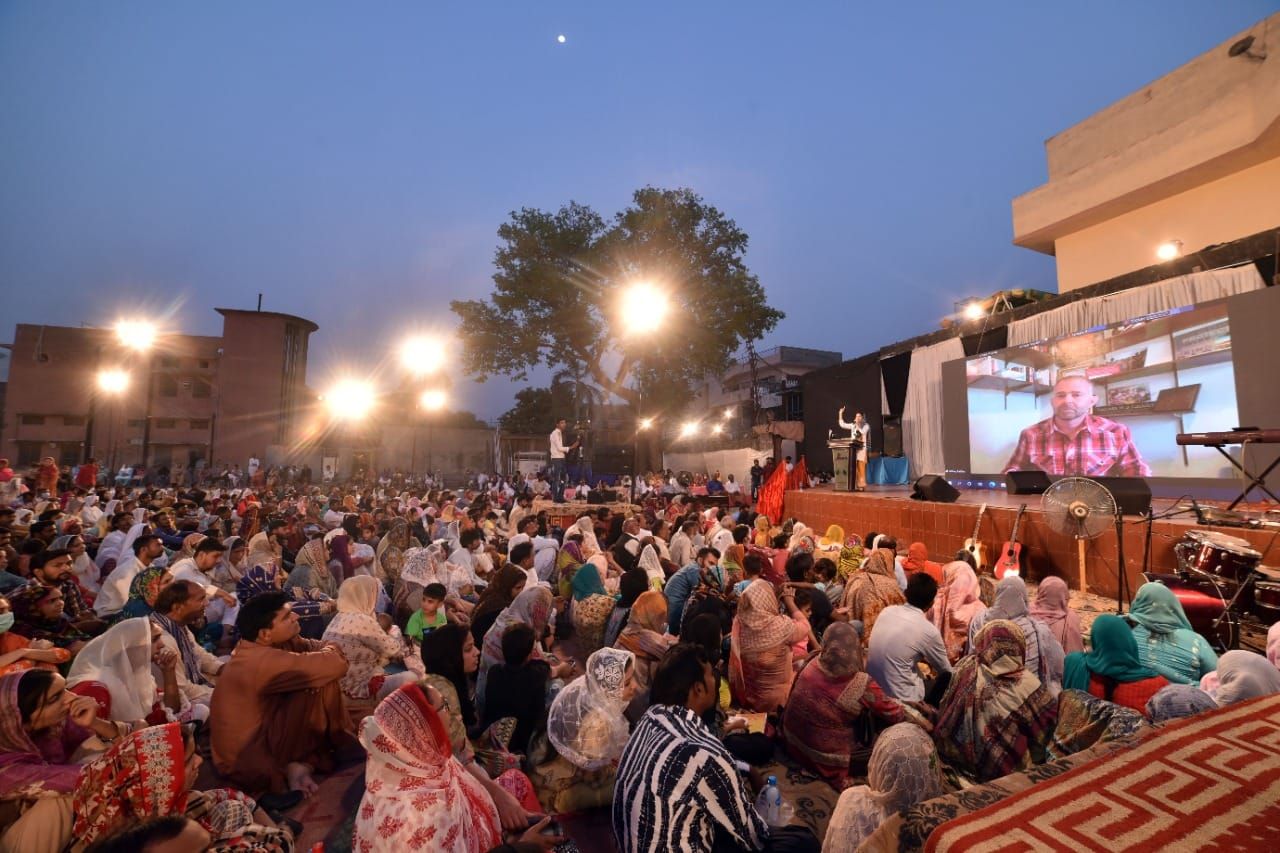 Large outdoor gathering watching a projected screen, likely an event or religious service. Many people seated.
