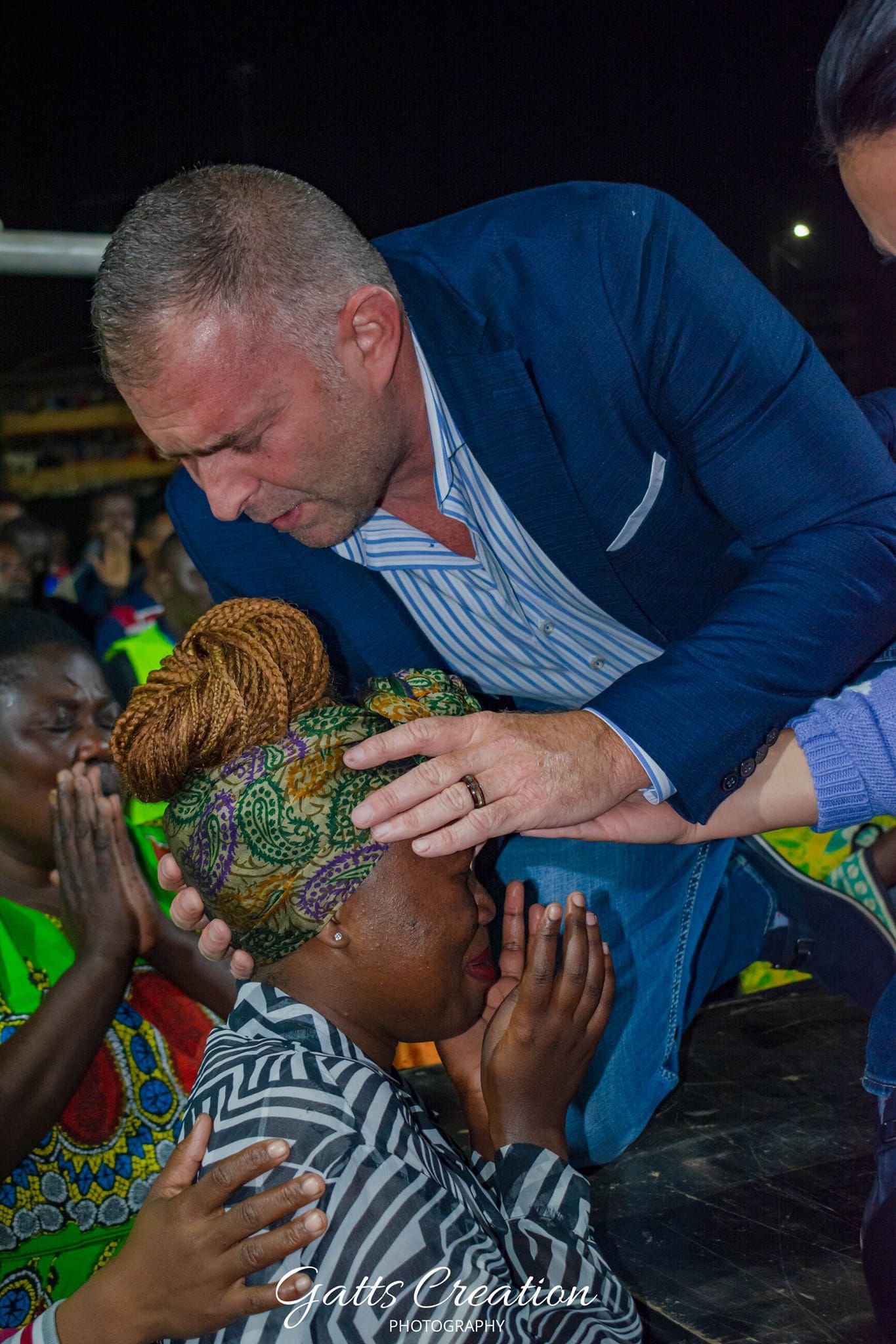 Man in blue jacket laying hands on woman's head; people praying.