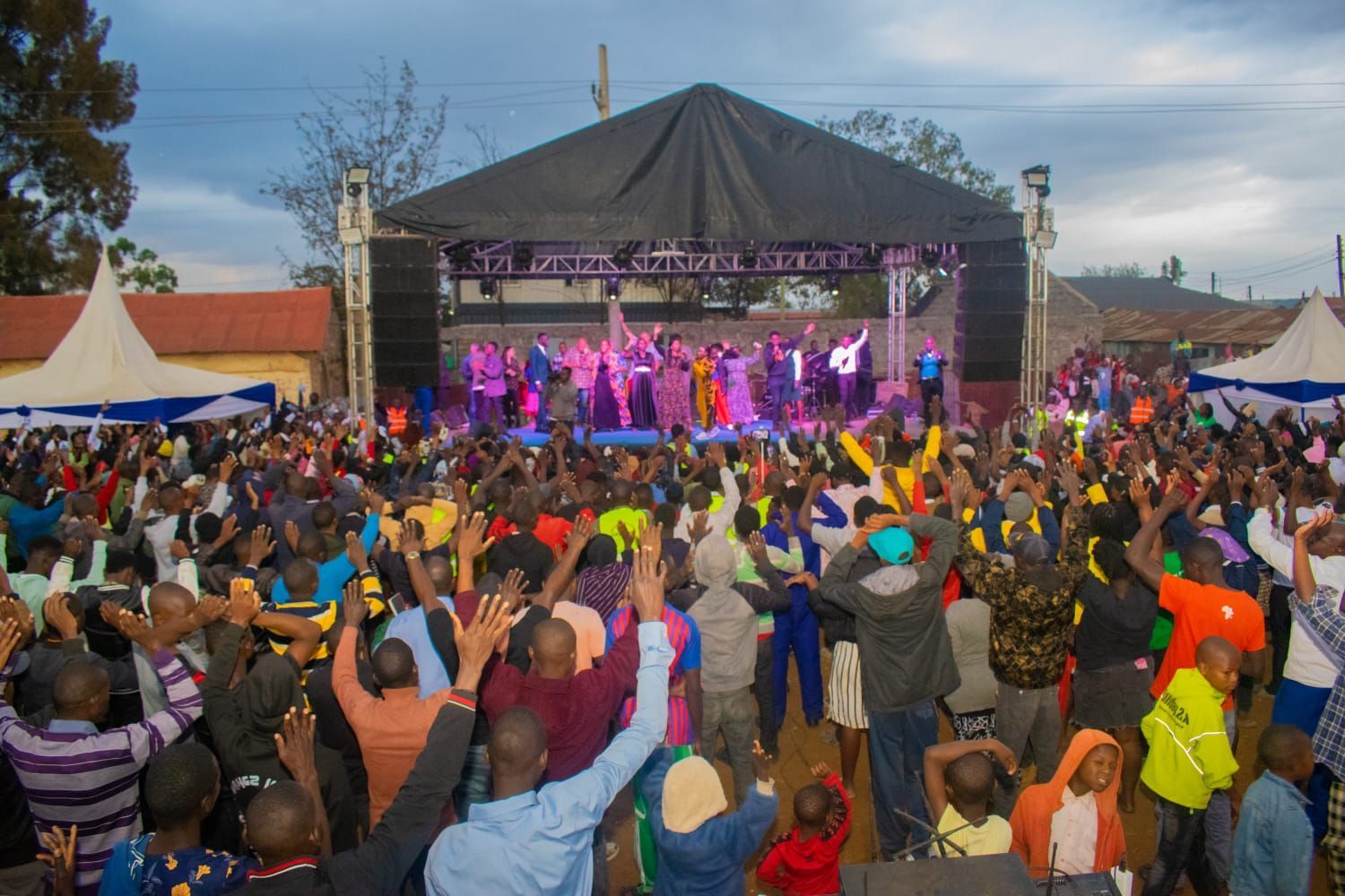 Large crowd at an outdoor concert, hands raised towards stage, brightly lit performers.