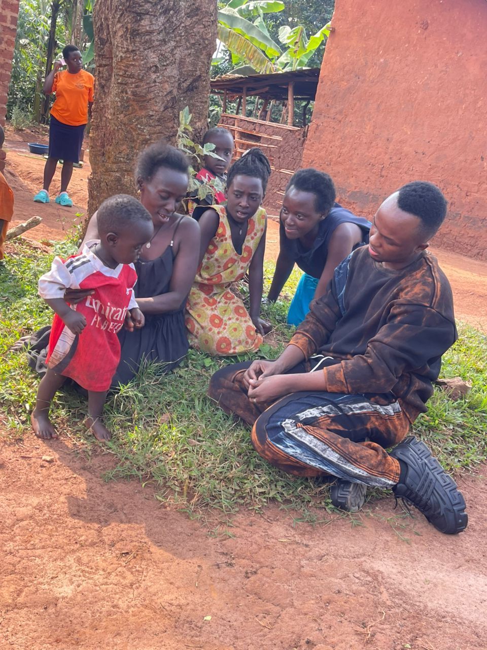 People gather around a child near a tree. One man kneels, talking to the child. Others watch nearby.