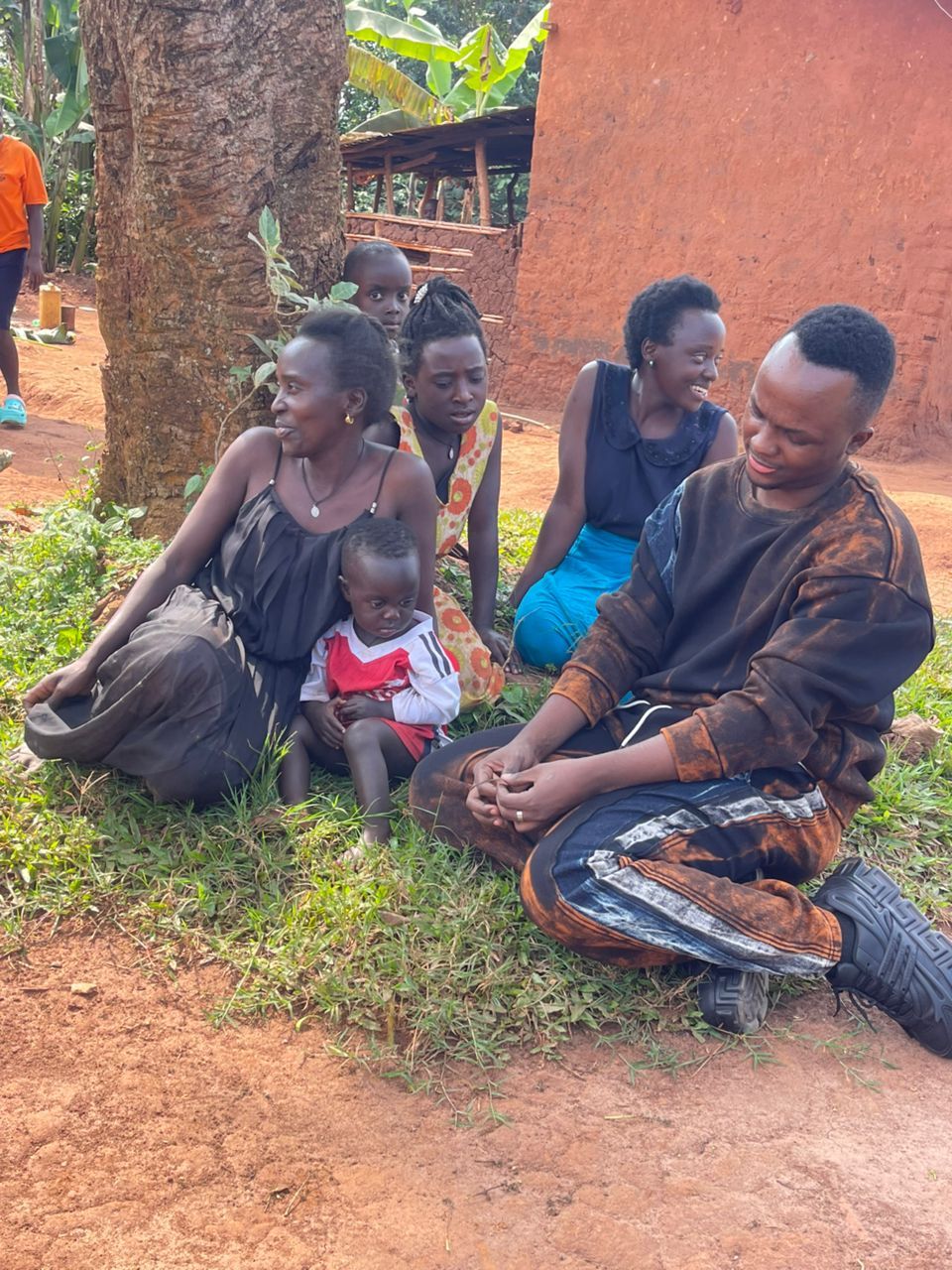 Group of people sitting outdoors on grass, some looking to the side. A child sits in front.