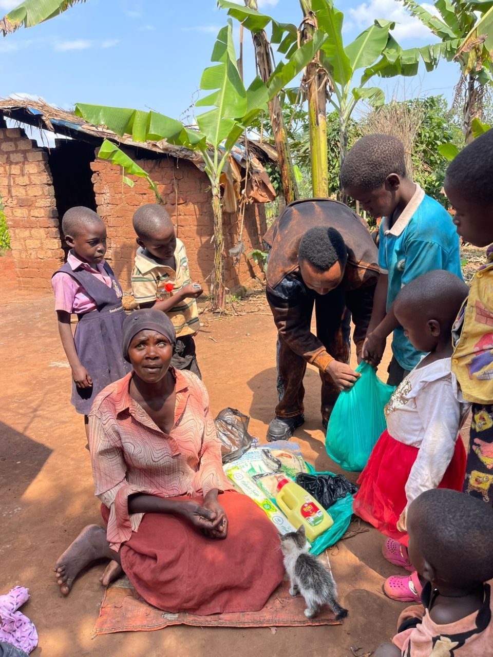 People gather around a seated woman receiving donations. Children observe as someone unpacks bags. Outdoors, simple buildings visible.