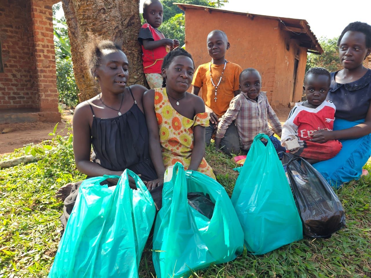 People with bags pose outdoors. Some sit and others stand near a mud-brick building.