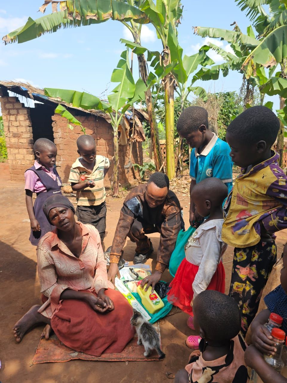 A man kneeling with a group of children and a woman in front of a house, possibly giving out goods.