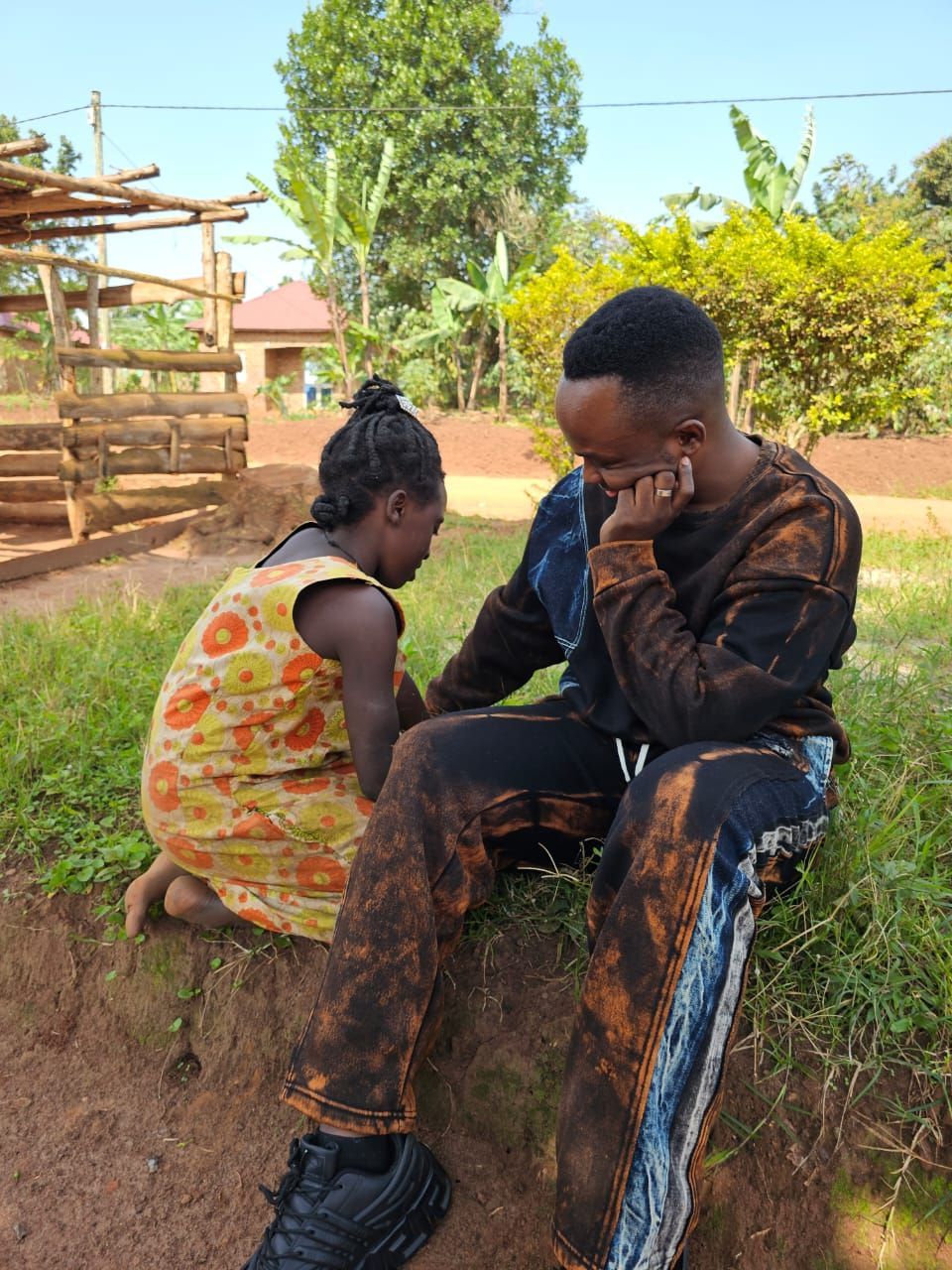 A person kneels near a seated person on grassy bank. They are in an outdoor setting.