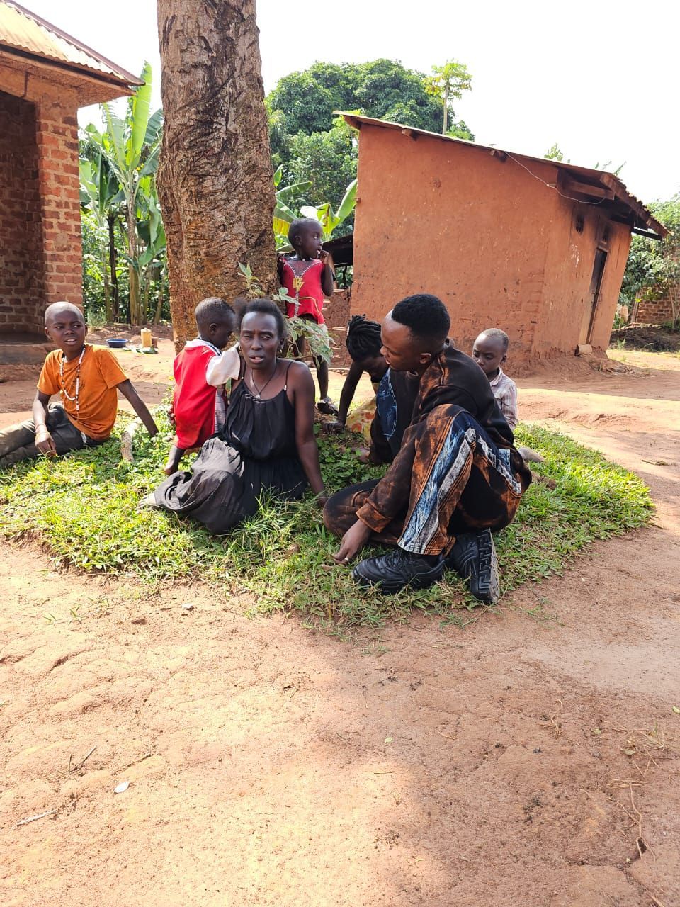 People, including children, sit near a tree and mud-brick structures in a grassy area.