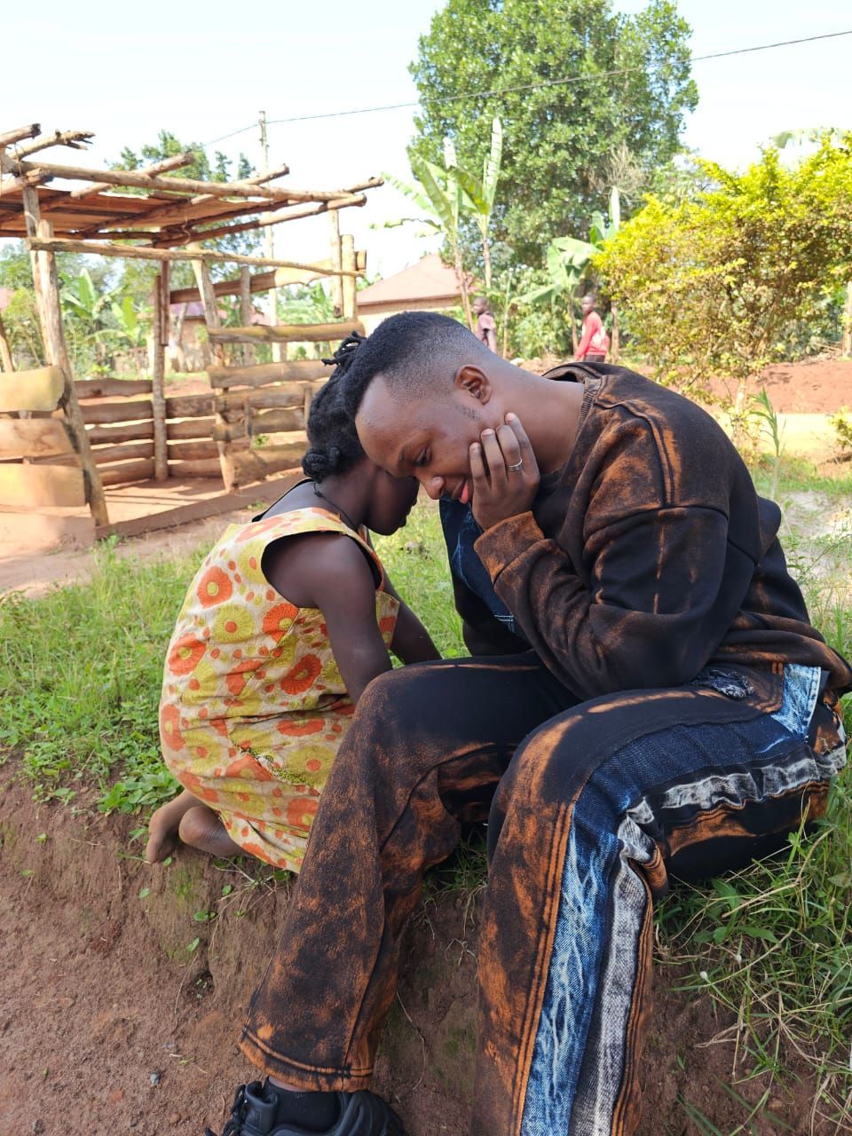 Two people sit outside; one leans on the other, both appear distressed. Earthy tones, overcast day.