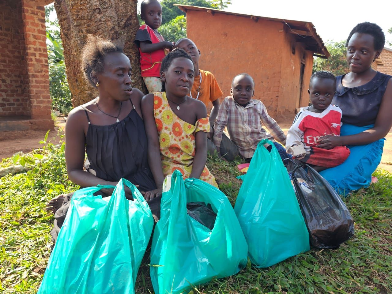 Family sitting on grass with bags of food near a tree and a small brick building.