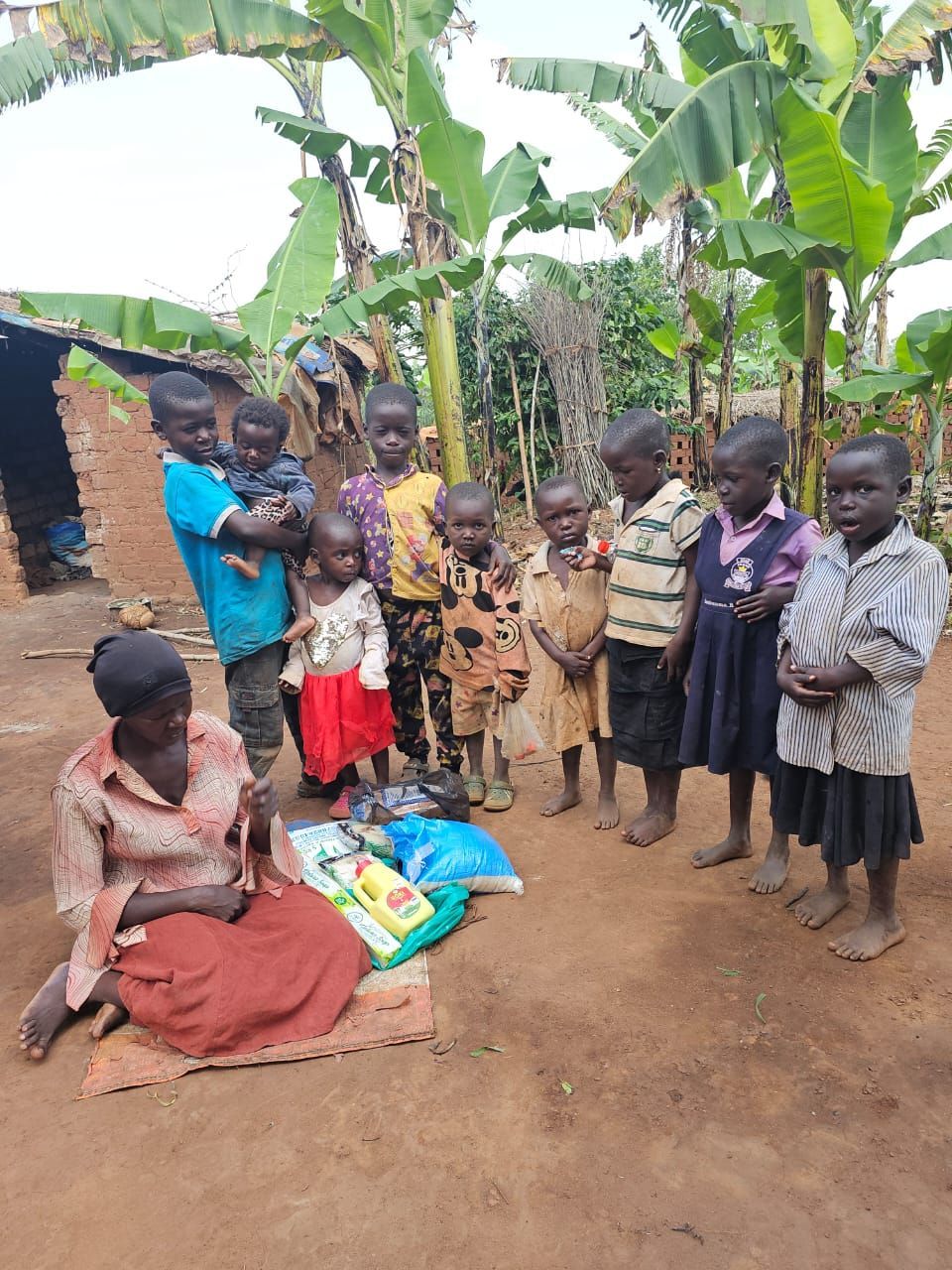 Woman and nine children in front of a dwelling. Children stand in a row; some are holding items.
