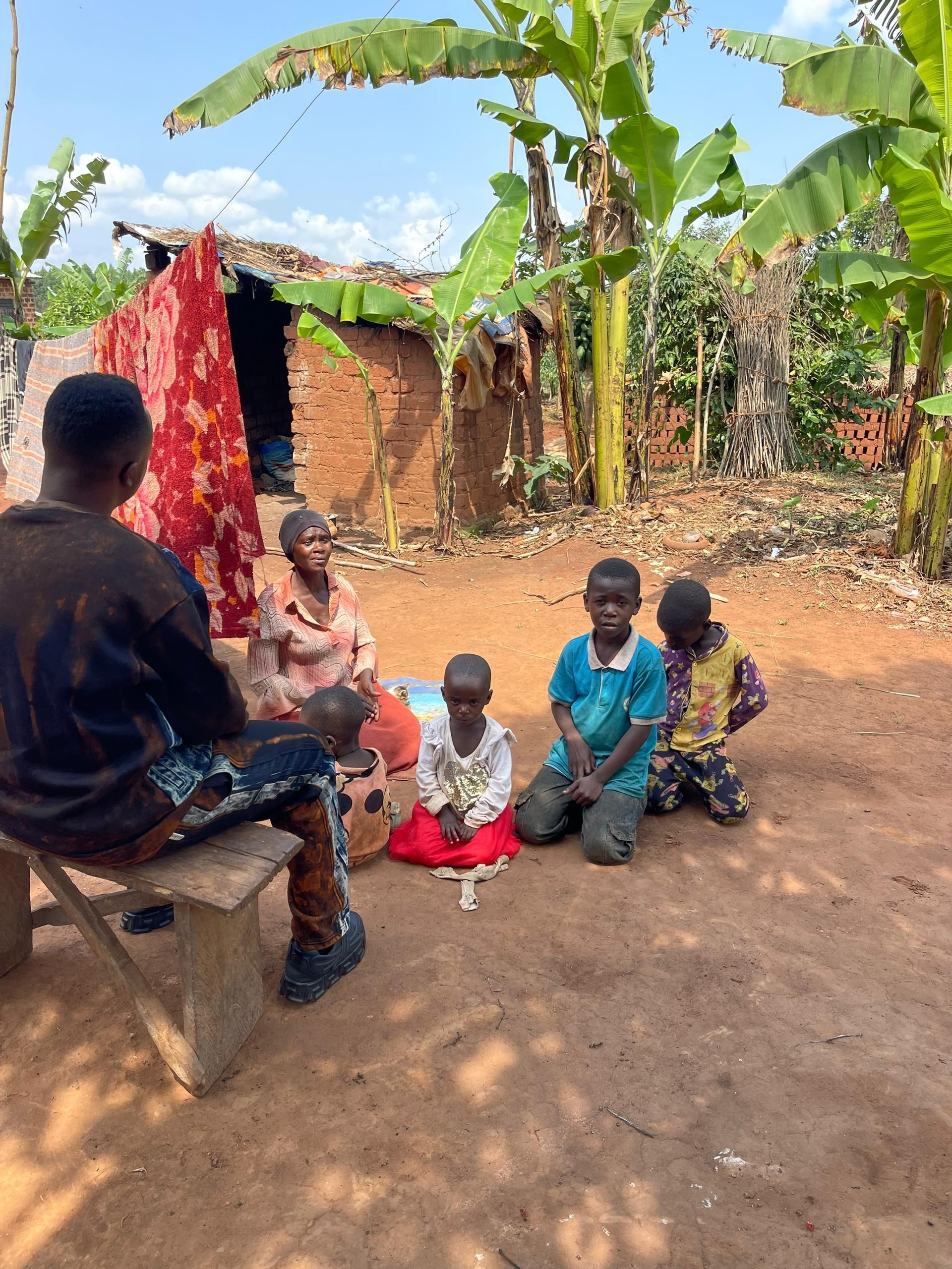 A person sits on a bench facing a group of children and an adult woman in front of a rustic building.