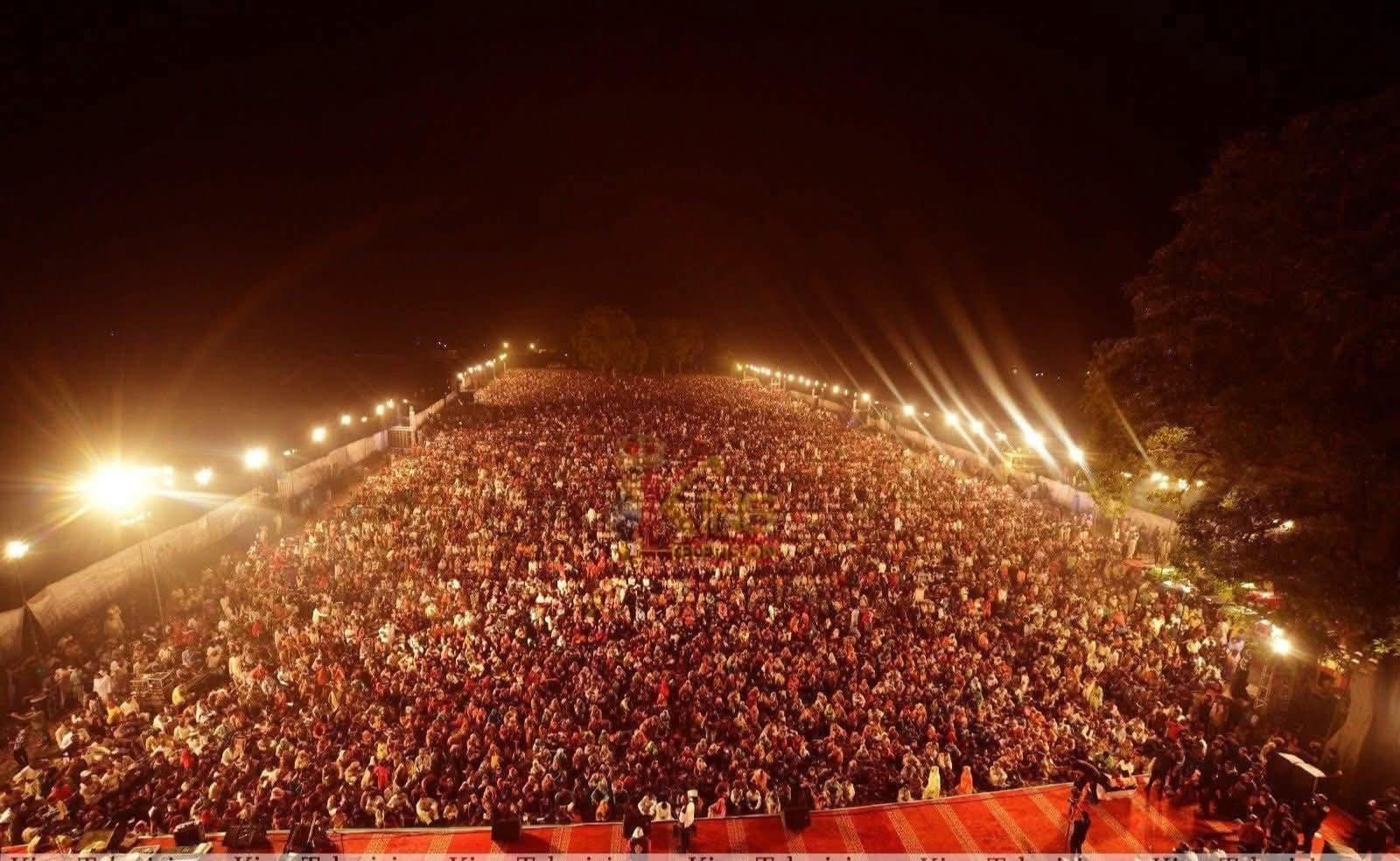 Large crowd gathered at night, illuminated by lights, likely an outdoor event.