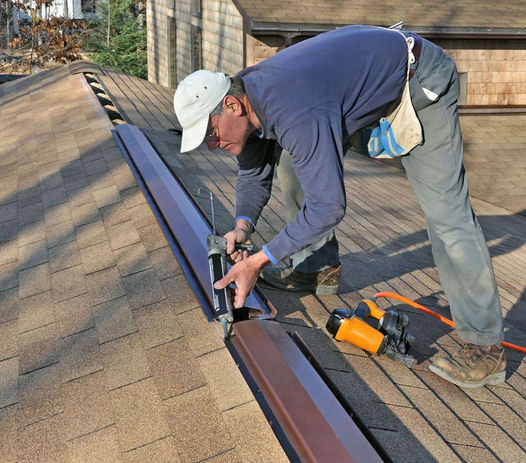 Man installing metal flashing on a roof with a nail gun. Brown roof, blue shirt, white hat.