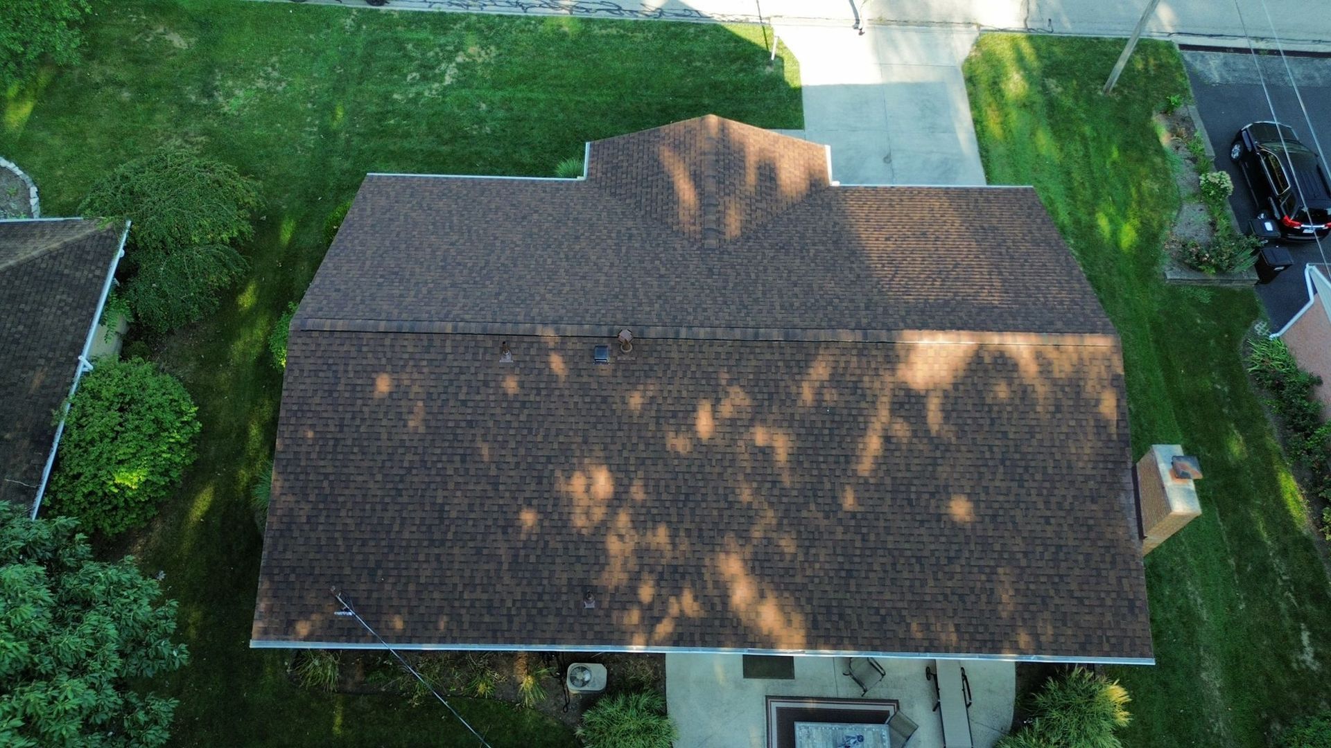 Aerial view of a brown shingled roof casting shadows, surrounded by green grass and a driveway.