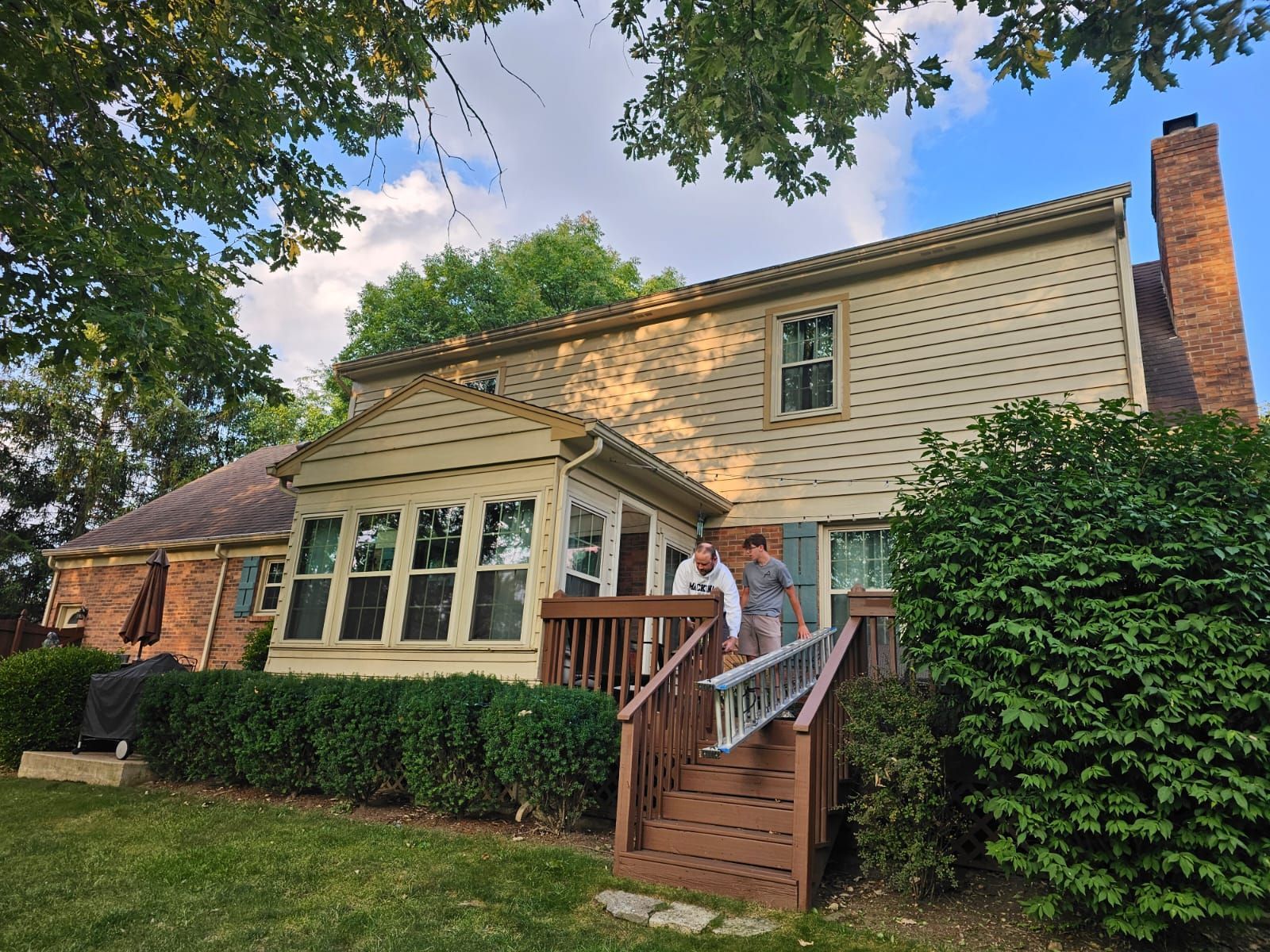 Backyard view of a two-story beige house with a brick chimney and deck; two people carrying a ladder.