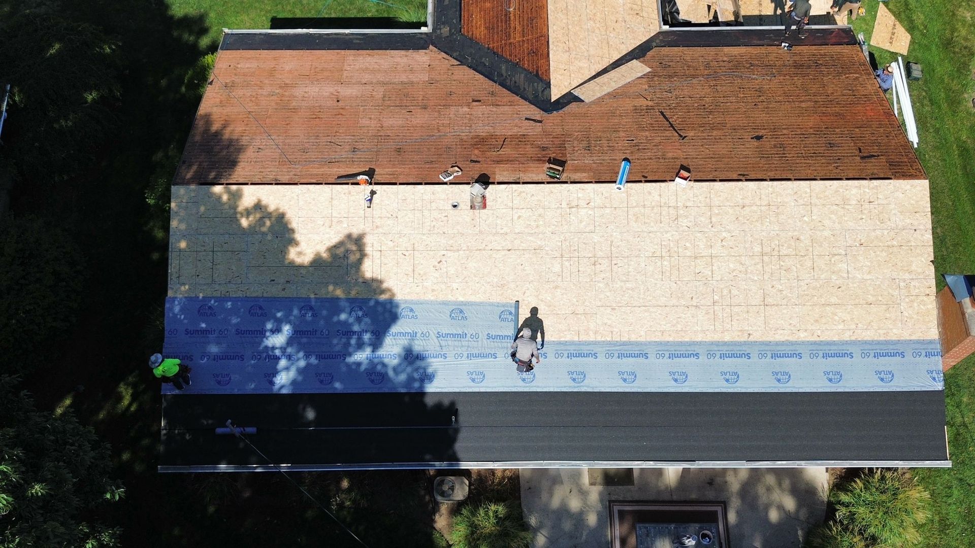 Aerial view of roof repair: workers applying roofing materials to a house. Different stages of the roof's renovation are visible.