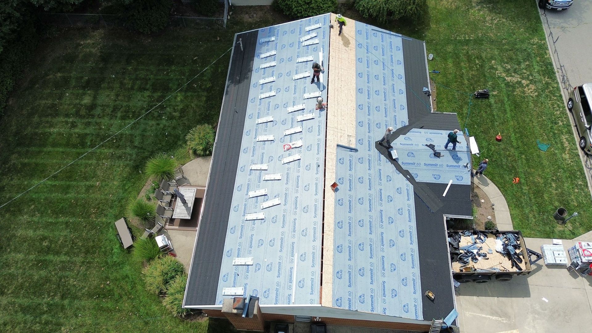 Roofing crew working on a house roof, using tools and materials; sunny day, green grass.
