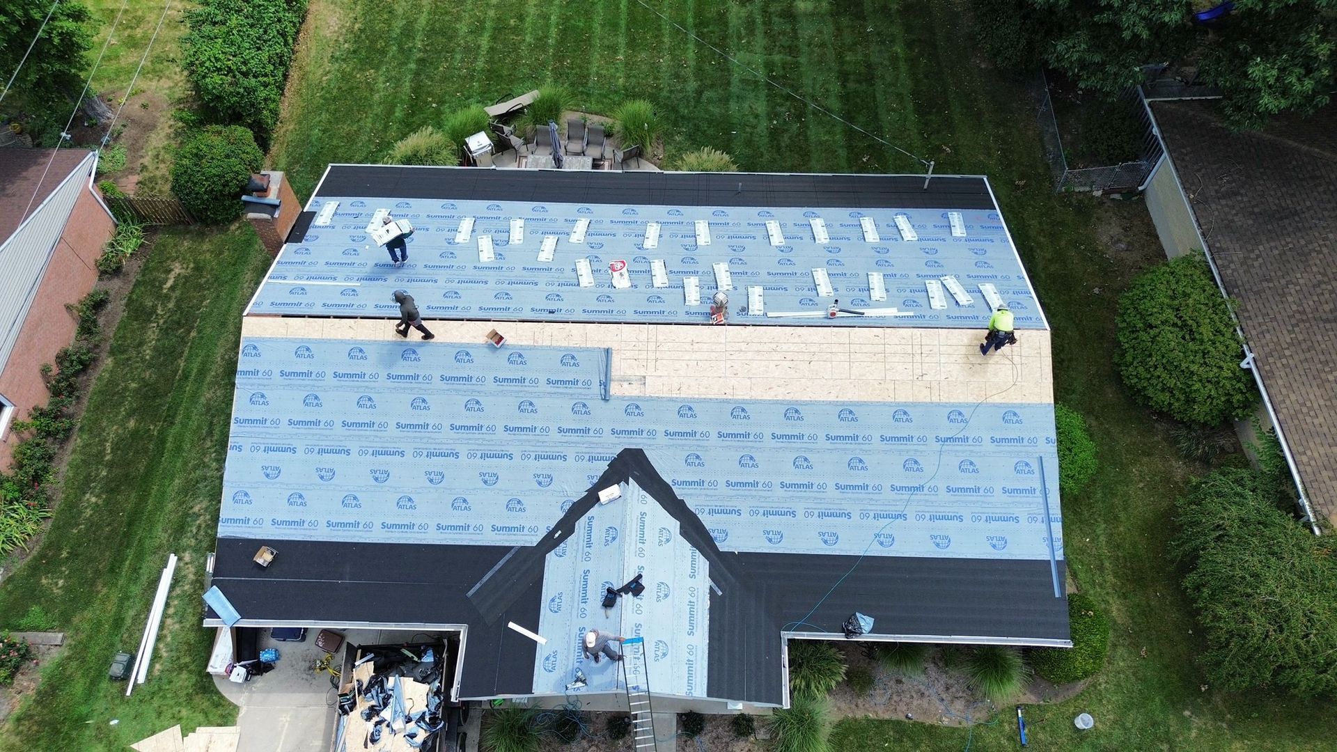 Aerial view of a roof being shingled by several workers. Blue underlayment covers most of the roof.