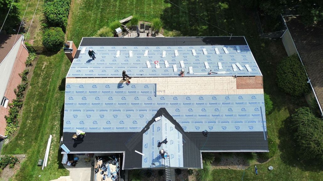 Roofing workers install a new roof on a house, placing blue underlayment and shingles.