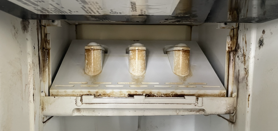Inside of a refrigerator, a white shelf holds three brown water filters with staining on the surrounding walls.