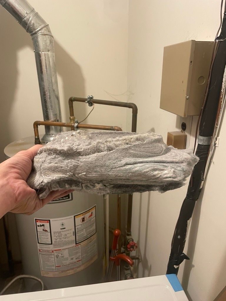 Hand holding gray insulation near a hot water heater. Copper pipes, electrical box, and metal ductwork visible.