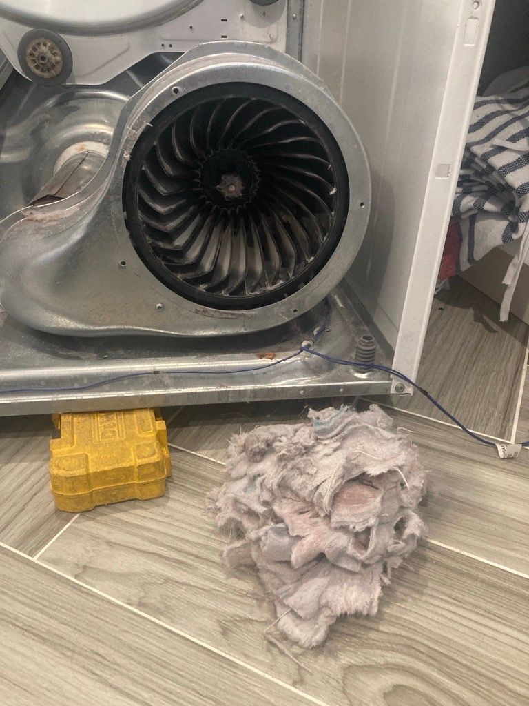 Open dryer showing blower fan with lint and a pile of lint on the floor.