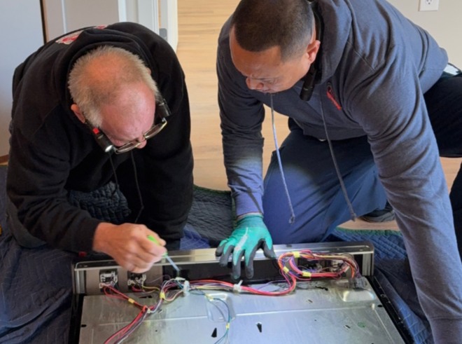 Two people kneeling, inspecting the wiring on the underside of a stovetop.