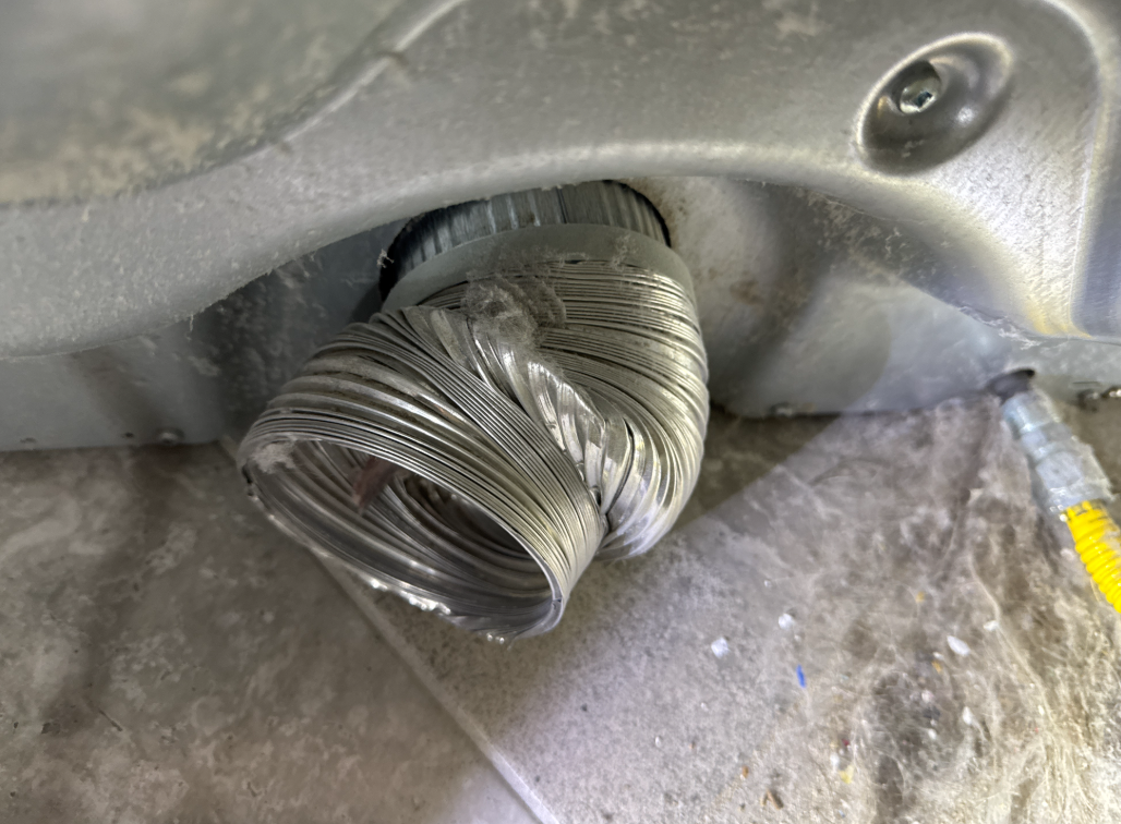 A crushed, silver dryer vent hose connected to a dryer. The setting is dusty.