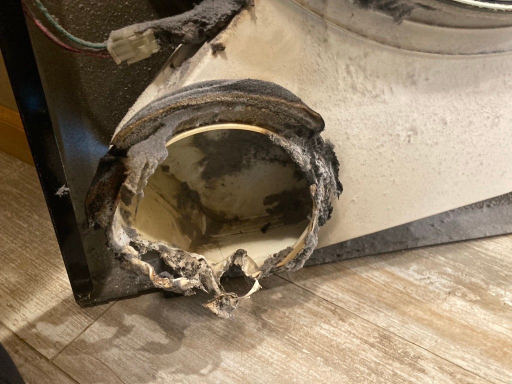 Damaged dryer vent opening with charring and debris, resting on a wood floor.