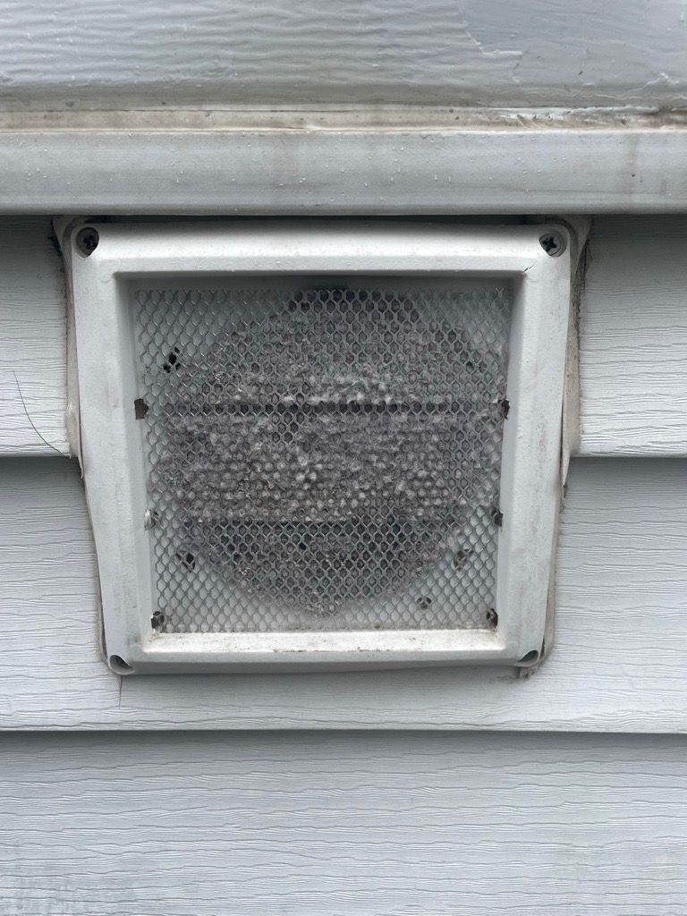 A rectangular, white dryer vent on gray siding is covered with lint.