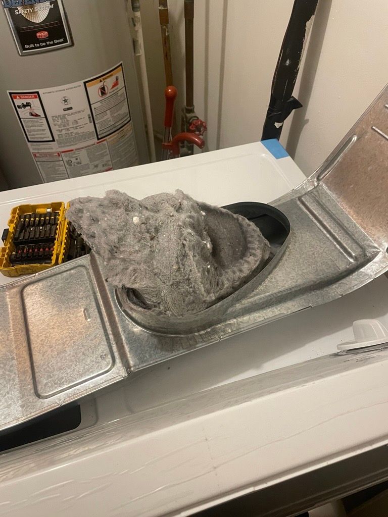 Lint-filled dryer vent, opened on top of a white washing machine. A screwdriver set sits to the left.