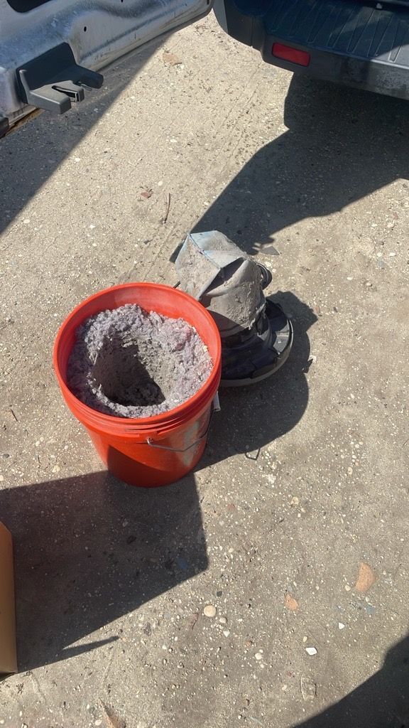 Red bucket filled with grey insulation on concrete, near damaged metal object. Outdoors.