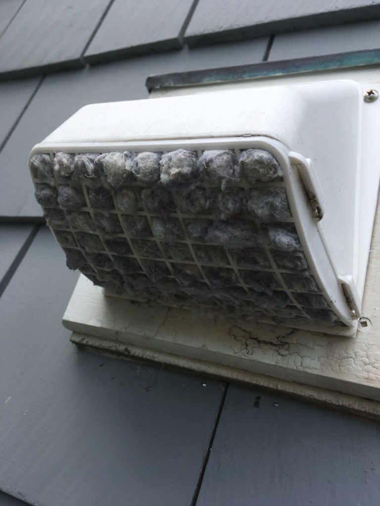 White vent cover on a gray roof, with a clogged, gray, grid-covered opening.